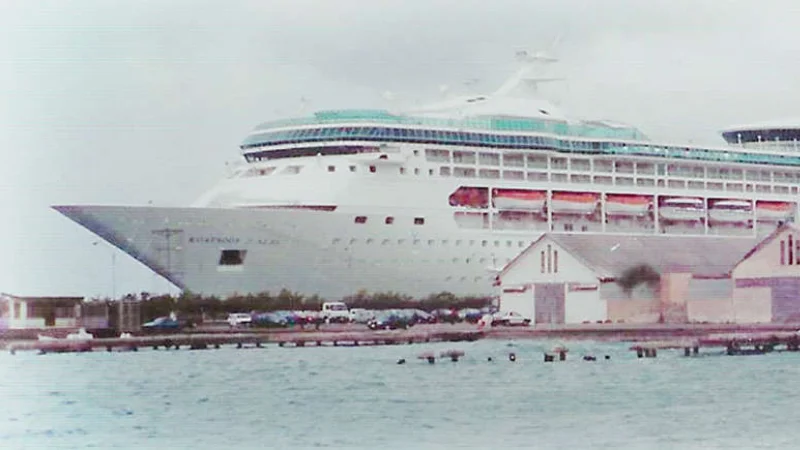 Rhapsody of the Seas docked at port, viewed from the shoreline on a cloudy day.